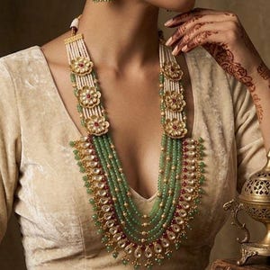 Peut inclure: Un collier orné et des boucles d'oreilles assorties. Le collier présente plusieurs rangées de perles vertes, entrecoupées d'éléments floraux dorés et d'accents rouges. Les boucles d'oreilles reprennent le motif floral. Les bijoux sont portés avec un haut crème.