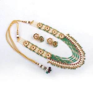K&ouml;nnte beinhalten: Ein verziertes Halsketten- und Ohrring-Set. Die Halskette hat mehrere Str&auml;nge aus gr&uuml;nen Perlen, die mit wei&szlig;en und goldenen Blumendesigns und roten Akzenten versehen sind. Die passenden Ohrringe haben ein &auml;hnliches Blumendesign. Das Set wird auf wei&szlig;em Hintergrund pr&auml;sentiert.