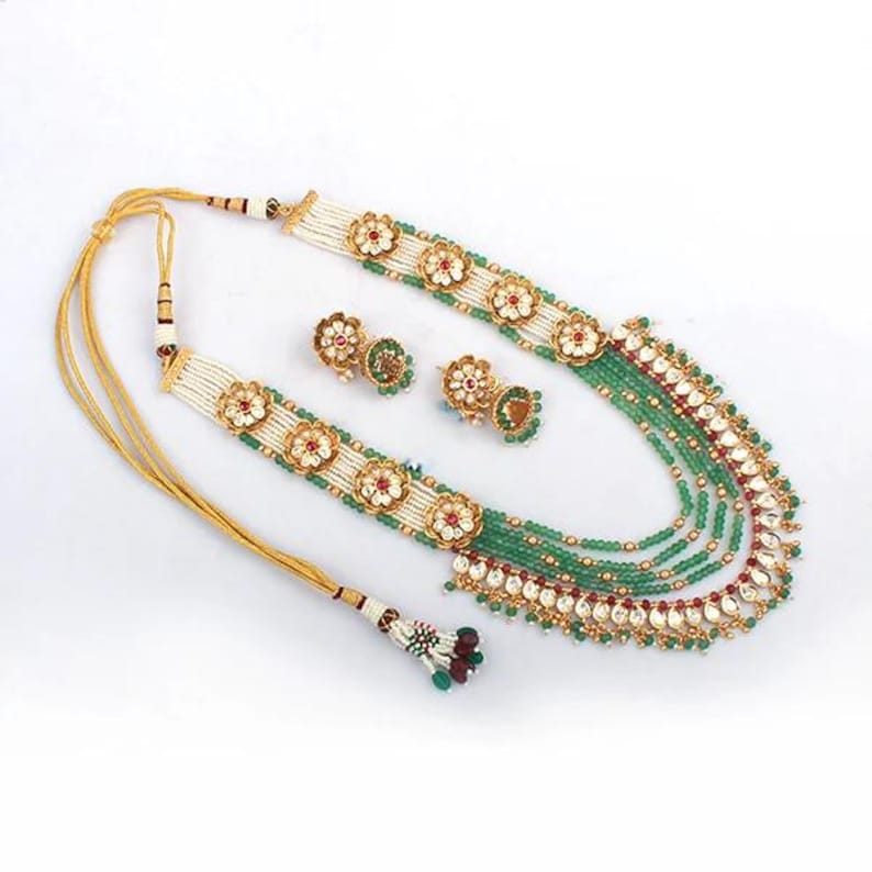 K&ouml;nnte beinhalten: Ein verziertes Halsketten- und Ohrring-Set. Die Halskette hat mehrere Str&auml;nge aus gr&uuml;nen Perlen, die mit wei&szlig;en und goldenen Blumendesigns und roten Akzenten versehen sind. Die passenden Ohrringe haben ein &auml;hnliches Blumendesign. Das Set wird auf wei&szlig;em Hintergrund pr&auml;sentiert.