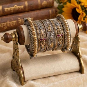 Op de afbeelding: Een set van vier versierde metalen armbanden op een standaard van hout en stof. De armbanden hebben ingewikkelde texturen, goudkleurige kralenranden en kleine rode steentjes. De standaard heeft een horizontale, met stof beklede cilinder.