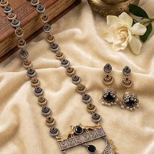 Könnte beinhalten: Ein langes Halsketten- und Jhumka-Ohrring-Set mit abwechselnden silber- und goldfarbenen kreisförmigen Gliedern. Der Anhänger zeigt zwei Pfauenfiguren, schwarze Steine und hängende weiße Perlen. Die Ohrringe sind mit schwarzen Steinen besetzt.