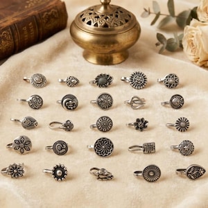 Peut inclure: Une collection de vingt-cinq anneaux de nez argentés disposés en cinq rangées. Chaque pièce présente un design circulaire ou géométrique unique avec des motifs en relief complexes, incluant des fleurs, des soleils et des formes abstraites.