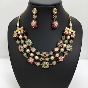 May include: A three-strand necklace and matching earrings set. The necklace features burgundy, gold, and pearl-coloured beads with floral and geometric accents. The earrings have a similar design, with burgundy and gold elements.