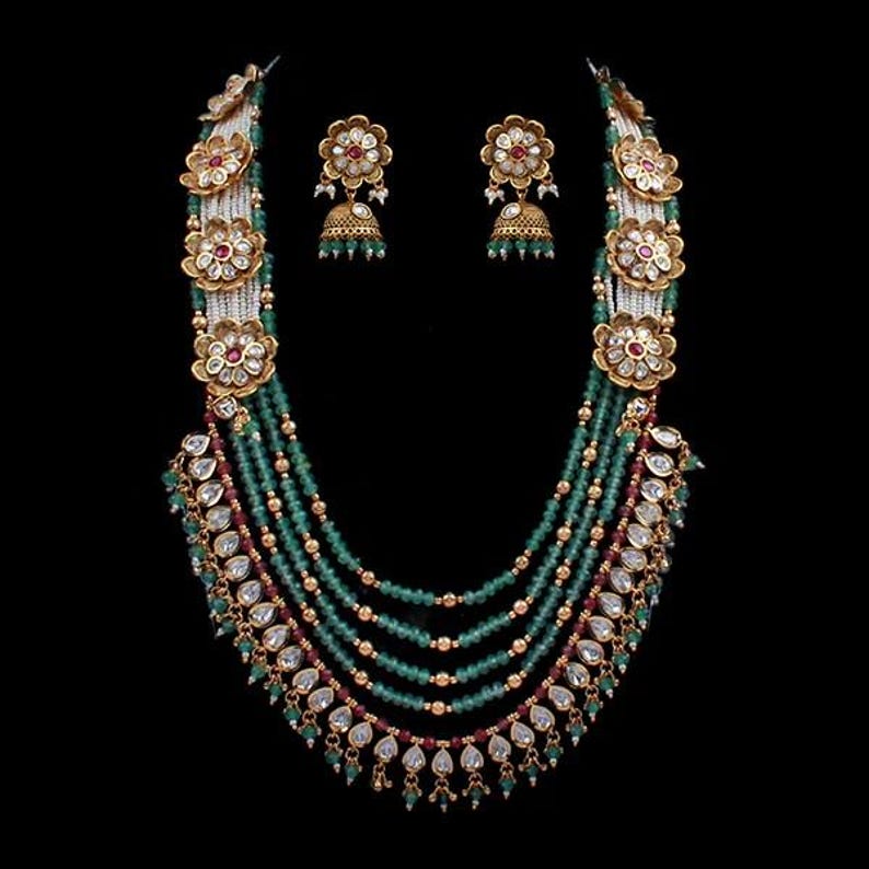 K&ouml;nnte beinhalten: Ein aufwendiges Halsketten- und Ohrring-Set. Die Halskette hat mehrere Str&auml;nge aus gr&uuml;nen Perlen, goldfarbene Details und florale Anh&auml;nger mit roten und wei&szlig;en Steinen. Die Ohrringe haben ein passendes florales Design mit gr&uuml;nen glockenf&ouml;rmigen Anh&auml;ngern.