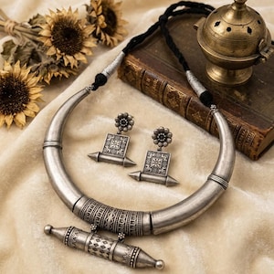 Pu&ograve; includere: Una collana in metallo color argento con un design a girocollo spesso e curvo con un pendente cilindrico centrale, abbinata a orecchini pendenti quadrati coordinati. Il set include un cordino nero con dettagli color argento e incisioni.