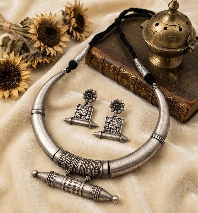 Pu&ograve; includere: Una collana in metallo color argento con un design a girocollo spesso e curvo con un pendente cilindrico centrale, abbinata a orecchini pendenti quadrati coordinati. Il set include un cordino nero con dettagli color argento e incisioni.