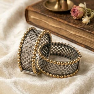 May include: Two wide bangles featuring a silver-toned mesh center with a repeating circular pattern, bordered by rows of small, polished gold-toned beads along the top and bottom edges. The bracelets have a structured, cylindrical shape.