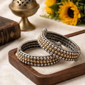 May include: Two hinged bangle bracelets featuring a multi-row design of small metallic spheres. The center row consists of gold-toned beads, flanked by two rows of silver-toned beads on each side. Each bracelet has a secure metal hinge mechanism.