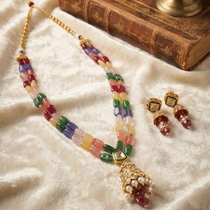 May include: A multi-strand necklace featuring polished beads in shades of red, green, blue, yellow, and pink, accented with a central gold-toned pendant with white pearls and a red stone, paired with matching drop earrings with floral patterns.