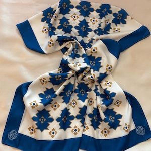 Op de afbeelding: Een witte sjaal met een herhalend patroon van blauwe stervormige vormen en kleine gouden vierkantjes. De sjaal heeft een blauwe rand en een decoratief embleem in de hoek. De sjaal is gevouwen en gedrapeerd.
