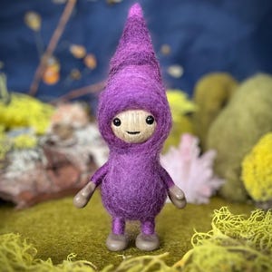 May include: A whimsical felted gnome figurine in shades of purple, with a wooden face and small gray hands and feet. The gnome wears a pointed hat and stands on a bed of green moss. The background is a blurred mix of greens and blues.