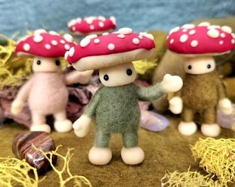 May include: A whimsical scene featuring four felted mushroom figures with red caps dotted with white spots. The figures have beige stems, white hands and feet, and are posed on a mossy surface. A few small stones and yellow moss are also visible.