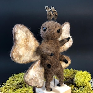May include: A brown felt moth with large wings, standing on a wooden base. The moth has black eyes and a pair of small, brown wings on its head.