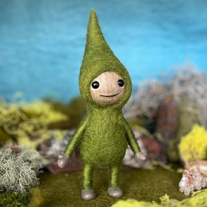 May include: A handmade felt doll with a green hooded outfit and a wooden face. The doll has a pointed hat, small arms and legs, and a smiling expression. The background features moss and a blue sky.