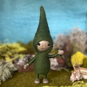 May include: A handcrafted felt doll with a whimsical design. The doll features a green hooded outfit and a wooden face with a smiling expression. The doll's arms and legs are also made of wood. The background includes moss and a blue sky.