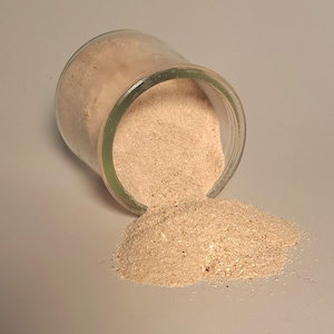 May include: A clear glass jar tipped over, spilling a light brown powder onto a white surface. The powder appears granular and has a slightly beige hue. The jar has a green rim.