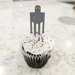 Puede incluir: Un cupcake de chocolate con glaseado blanco y chispas de chocolate cubierto con un robot gris como adorno de cupcake con ojos amarillos.