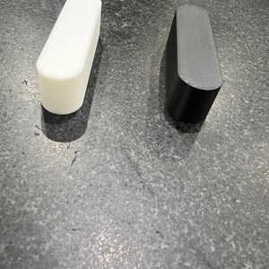 May include: Two rectangular objects with rounded edges. One is white, the other is black. They stand upright on a grey surface. Both have a smooth finish. The objects are similar in shape and size, suggesting they are a pair.