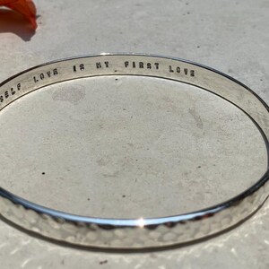 May include: Silver hammered bangle bracelet with the inscription "Self Love is my first love".