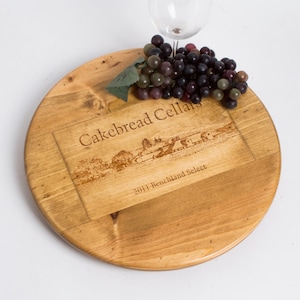 May include: A round wooden serving board with the text "Cakebread Cellars" and "2011 Benchland Select" etched into the wood. A bunch of grapes and a wine glass are on the board. The board is light brown.