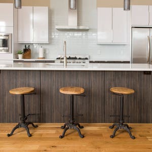 May include: Three wooden and metal bar stools with black metal frames and round wooden seats. The stools are in front of a dark wood kitchen island with a white countertop.