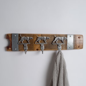 May include: A wooden wall-mounted coat rack with three hooks shaped like hunting dogs. The rack is made from reclaimed wood and has a rustic finish. A gray knitted scarf is hanging from one of the hooks.