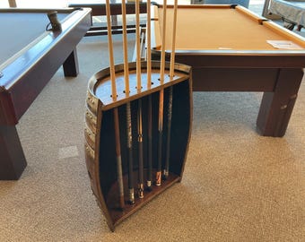 May include: A floor-standing pool cue rack crafted from a repurposed wooden barrel. The dark brown curved wooden frame features a flat top with five circular cutouts holding five individual pool cues in a vertical, upright position.