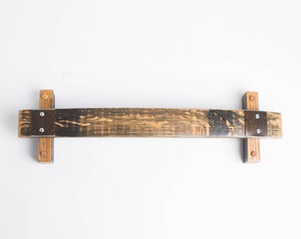 May include: A wooden shelf made from a reclaimed wine barrel stave. The shelf is stained dark brown and has metal brackets on each end. The shelf is mounted to a white wall.