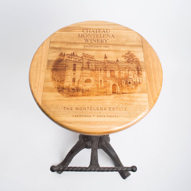 May include: A wooden bar stool with a round, brown wooden seat featuring a laser-etched image of Chateau Montelena Winery. The seat is inscribed with "Chateau Montelena Winery, Established 1882, The Montelena Estate, Calistoga, Napa Valley". The stool has a black metal base with a footrest.