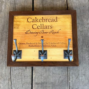 May include: A wooden sign with a brown frame and three hooks. The sign is engraved with the text "Cakebread Cellars Dancing Bear Ranch 2023 Grown, Produced & Bottled by Cakebread Cellars Rutherford, California, USA".