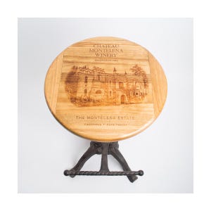Puede incluir: Un taburete redondo de madera con base de metal oscuro y reposapiés. El asiento presenta una imagen grabada con láser de un edificio de piedra y el texto CHATEAU MONTELENA WINERY ESTABLISHED 1882 THE MONTELENA ESTATE CALISTOGA NAPA VALLEY en negro.
