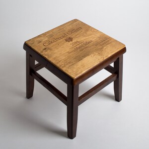 May include: A wooden stool with a dark brown frame and a square top made from a repurposed wine barrel. The top features a label that reads "Cakebread Cellars 2011  Napa Valley  Cabernet Sauvignon  Benchland Select  66750 ML".