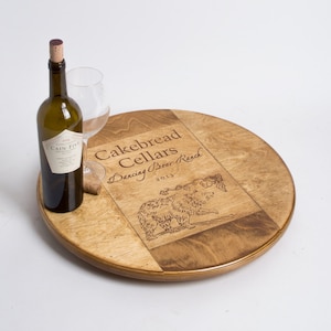 May include: A round wooden wine bottle holder with a laser engraved design featuring the text "Cakebread Cellars Dancing Bear Ranch 2013" and a bear illustration. A bottle of wine and a wine glass are on the holder.