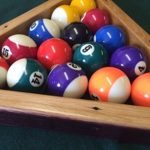 May include: A wooden triangle rack filled with billiard balls, including a solid red ball with the number 15, a solid green ball with the number 14, a solid purple ball with the number 4, and a solid orange ball with the number 1.