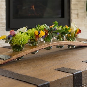 May include: A wooden wine barrel stave centerpiece with six small glass bowls, each holding a different arrangement of colorful flowers. The centerpiece sits on a wooden table with metal accents, in front of a fireplace.