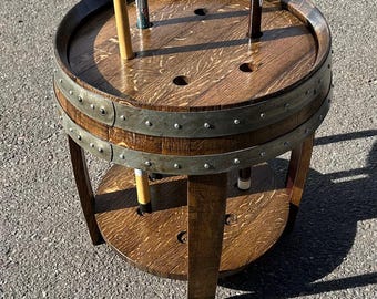 May include: A circular wooden pool cue rack constructed from a repurposed barrel, featuring a dark brown stained finish with visible wood grain. The rack includes two metal bands with rivets and multiple circular cutouts for holding pool cues.