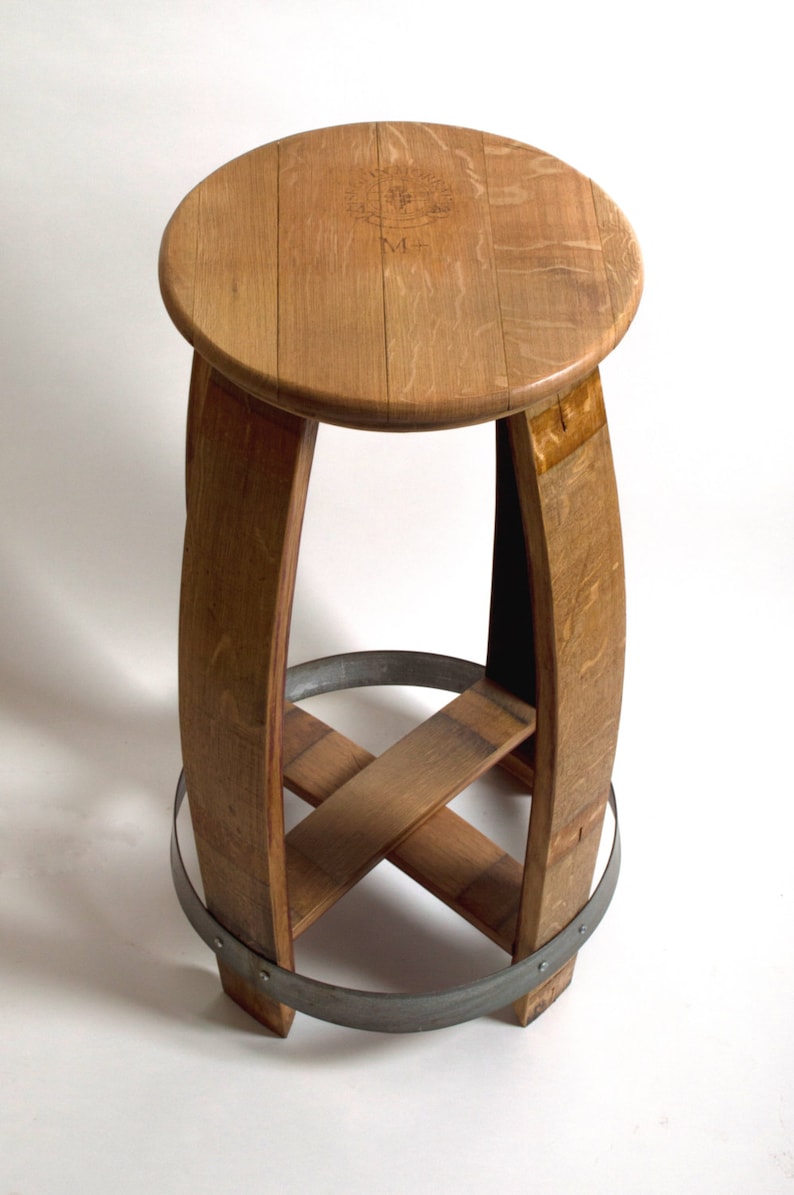 Puede incluir: Un taburete de madera hecho de un barril de vino reutilizado. El taburete tiene un asiento redondo y una banda de metal alrededor de la parte inferior.