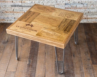 Puede incluir: Una mesa de café cuadrada de madera con una tapa hecha de madera de caja de vino reutilizada. La mesa tiene cuatro patas de horquilla y presenta una variedad de etiquetas de vino francés impresas en la tapa.