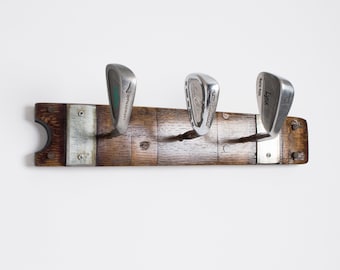 May include: A wooden wall-mounted coat rack made from a repurposed wine barrel stave. Three golf clubs are used as hooks. The clubs are a green and silver "Tourcraft" wedge, a silver "Callaway" wedge, and a silver "Lynx" wedge.