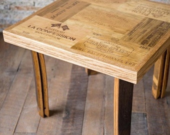 Puede incluir: Una mesa de café cuadrada de madera con una tapa hecha de duelas de barril de vino reutilizadas. Las duelas están impresas con los nombres de diferentes vinos, incluyendo Chateau La Confession Saint Emilion, Chateau Chasse-Spleen Moulis y Louis Jadot. La mesa tiene cuatro patas hechas de barriles de vino.
