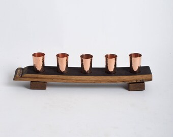 May include: Five copper shot glasses sit on a wooden stand made from a reclaimed barrel stave. The stand is dark brown with a natural wood grain.