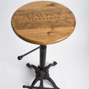 May include: A wooden bar stool with a round, brown wooden seat featuring the text "Caymus Vineyards Special Selection". The stool has a black metal base with a footrest.