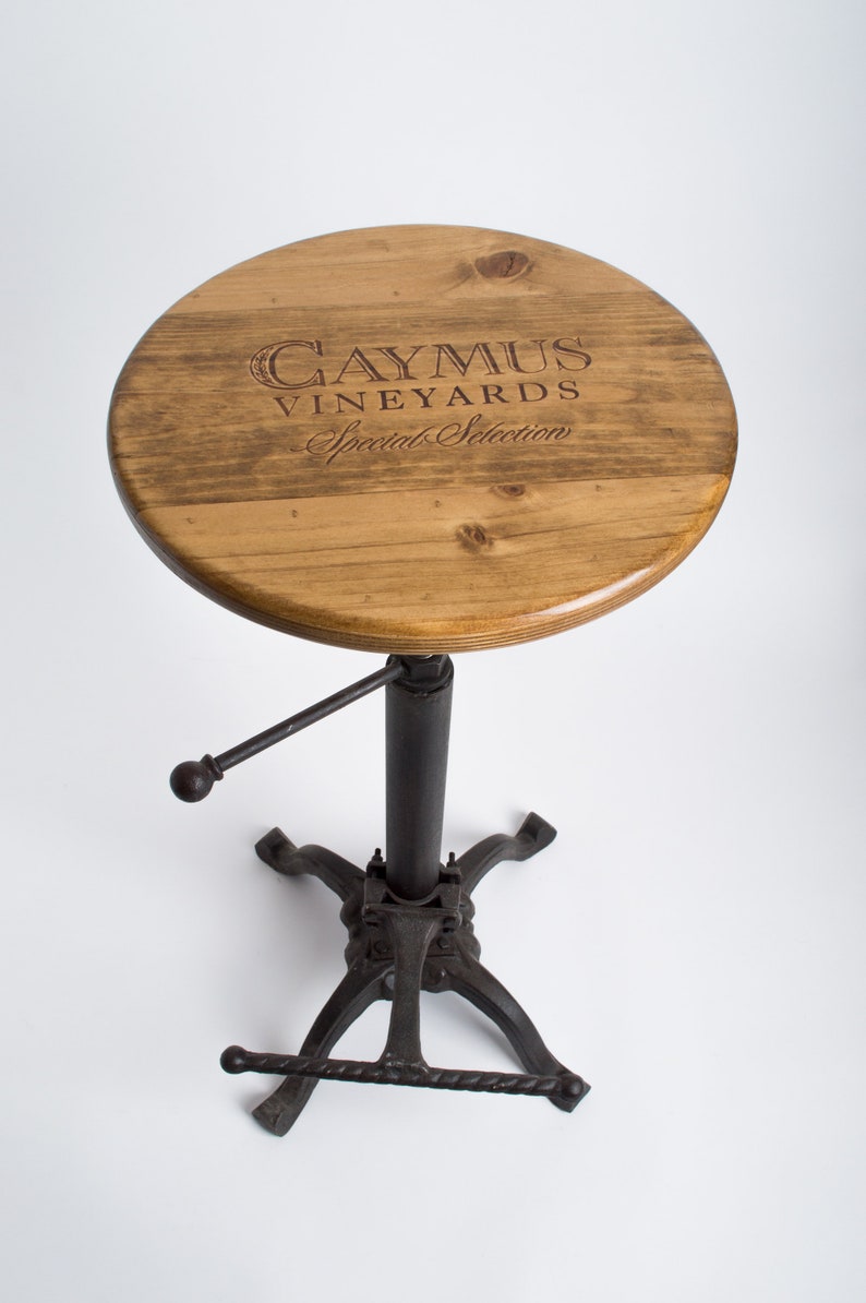 May include: A wooden bar stool with a round, brown wooden seat featuring the text "Caymus Vineyards Special Selection". The stool has a black metal base with a footrest.