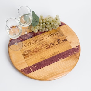 May include: A round wooden serving tray with a purple and brown inlay. The tray has a laser-etched design of a vineyard and the text "Cakebread Cellars 2008 Vine Hill Ranch". Two wine glasses filled with white wine and a bunch of green grapes are on the tray.