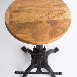 May include: A wooden stool with a round top and a black metal base. The top of the stool is made from a wine barrel stave and features the text "Opus 2004" and a signature. The base has a footrest and a lever for adjusting the height.