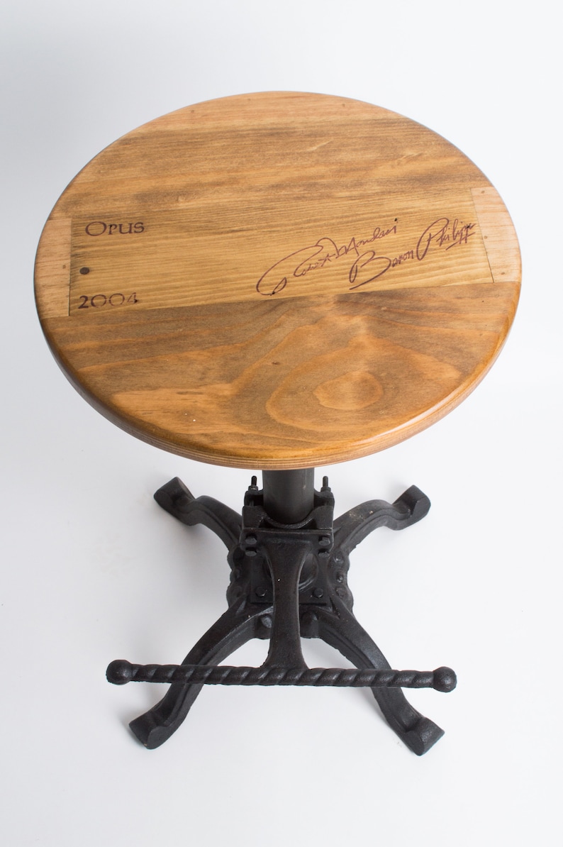 May include: A wooden stool with a round top and a black metal base. The top of the stool is made from a wine barrel stave and features the text "Opus 2004" and a signature. The base has a footrest and a lever for adjusting the height.