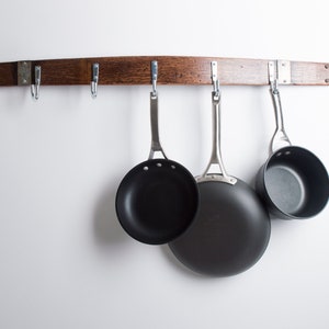 May include: A wooden wall-mounted rack with hooks for hanging kitchen utensils. Three black non-stick pans are hanging from the hooks.