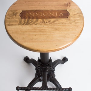May include: A wooden bar stool with a round, light brown wooden seat and a black metal base. The seat has the words "INSIGNIA 2012" engraved on it.