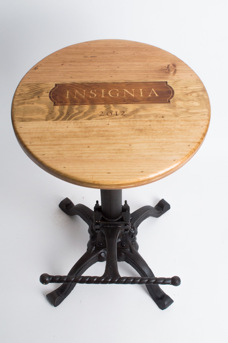 May include: A wooden bar stool with a round, light brown wooden seat and a black metal base. The seat has the words "INSIGNIA 2012" engraved on it.