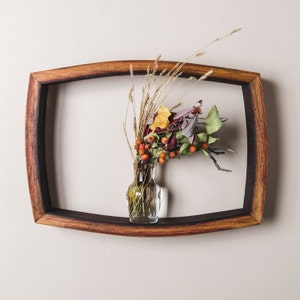 May include: A small glass vase with dried flowers and berries sits on a wooden shelf. The shelf is shaped like a half-circle and is made of dark wood.